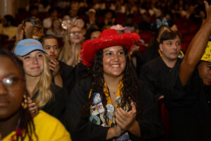 A student in a red cowboy hat and graduation robe and stole claps her hands 