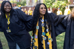 A student in a graduation robe and stole decorated with buttons smiles 