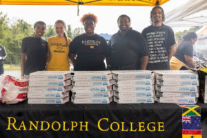 A group of people pose together behind a table with a Randolph College tablecloth with pizzas stacked on it 