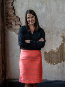A woman stands for a photo, with her arms crossed and a brick wall behind her 