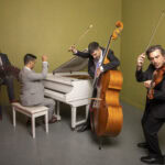A group of musicians in a room together. One sits at a piano, next to a man playing the accordian. Another man holds his cello, with one hand in the air holding the bow. Next to him is a violinist, also holding his bow in the air.