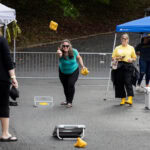 A woman throws a bean bag during a game of cornhole, while other participants look on