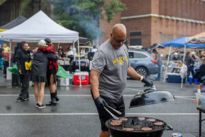 A man in a Randolph College shirt cooks hamburgers on a grill 