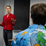 A woman stands in front of a classroom, speaking A woman stands in front of a classroom, speaking