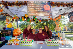 Four stduents in maroon shoots smile together in a fall festival booth
