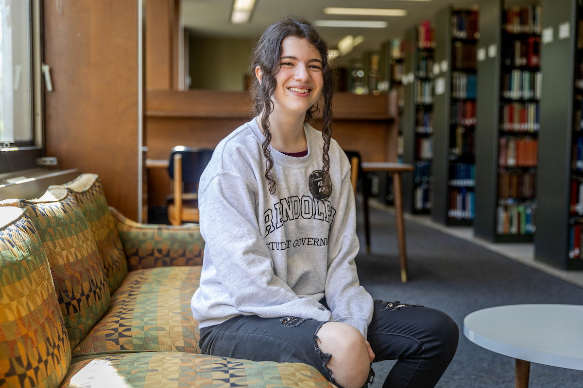 A student sits on a couch in the library, wearing a Randolph College sweatshirt