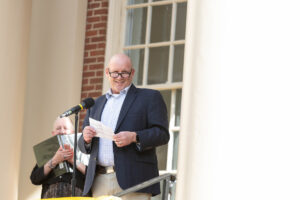 A man speaks in front of a mircophone, holding a piece of paper 