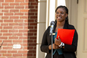 A female student stands in front of a microphone, holding a red folder.