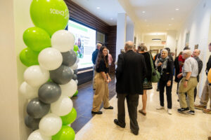 A group gathers inside a building, with green, white, and gray balloons and phots projected onto a screen in front of them 