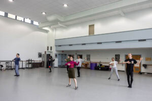 A woman in a skirt and magenta turtleneck demonstrates a dance move for gathered students in a dance studio
