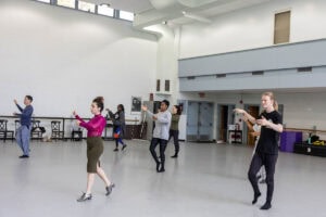 A woman in a skirt and magenta turtleneck demonstrates a dance move for gathered students in a dance studio