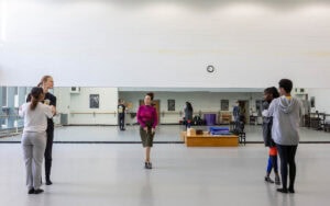 A woman in a skirt and magenta turtleneck demonstrates a dance move for gathered students in a dance studio