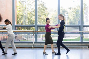 A woman in a skirt and magenta turtleneck demonstrates a dance move for gathered students in a dance studio