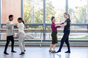 A woman in a skirt and magenta turtleneck demonstrates a dance move for gathered students in a dance studio
