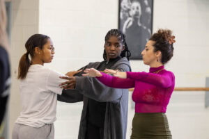 A woman in a skirt and magenta turtleneck demonstrates a dance move for gathered students in a dance studio