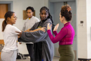 A woman in a skirt and magenta turtleneck demonstrates a dance move for gathered students in a dance studio
