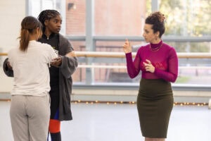 A woman in a skirt and magenta turtleneck demonstrates a dance move for gathered students in a dance studio