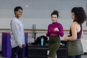 A woman in a skirt and magenta turtleneck demonstrates a dance move for gathered students in a dance studio