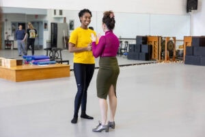 A woman in a skirt and magenta turtleneck demonstrates a dance move for gathered students in a dance studio