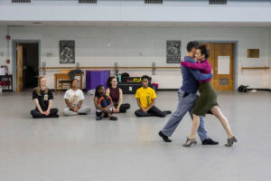 A woman in a skirt and magenta turtleneck demonstrates a dance move with a man in a blue shirt for gathered students in a dance studio