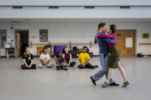 A woman in a skirt and magenta turtleneck demonstrates a dance move with a man in a blue shirt for gathered students in a dance studio