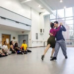 A woman in a skirt and magenta turtleneck demonstrates a dance move with a man in a blue shirt for gathered students in a dance studio A woman in a skirt and magenta turtleneck demonstrates a dance move with a man in a blue shirt for gathered students in a dance studio