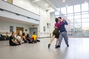 A woman in a skirt and magenta turtleneck demonstrates a dance move with a man in a blue shirt for gathered students in a dance studio