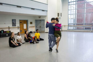 A woman in a skirt and magenta turtleneck demonstrates a dance move with a man in a blue shirt for gathered students in a dance studio