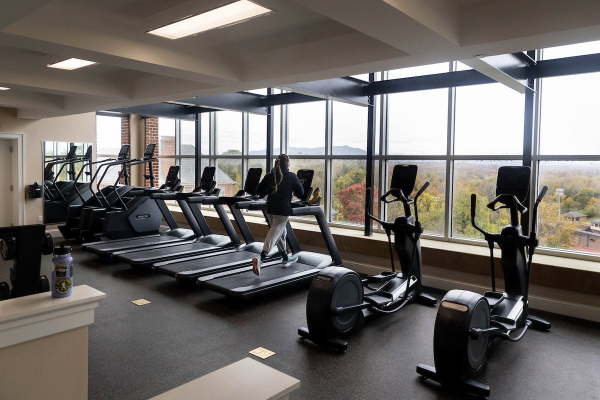 A student runs on a treadmill in a fitness center, facing a wall of windows with mountains int he distance 