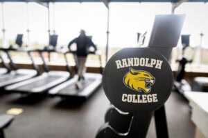 A close-up of a dumbell with the Randolph College WildCat head on it, with someone running on a treadmill in the background 