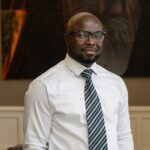 A man poses for a photo, wearing a tie and white shirt, with his hands in his pockets