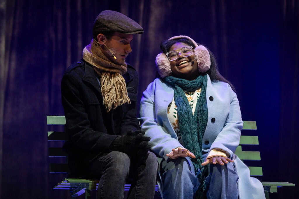 A man and a woman sit on a bench together onstage, wearing winter coats and earmuffs