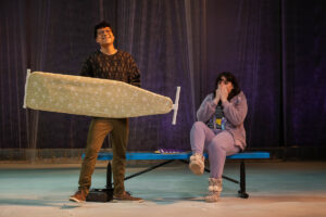 A man holds an ironing board in the air as a woman sitting nearby holds her hands over her mouth