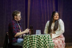Two actors sit onstage at a table, with a green and white checkered tablecloth, talking to each other 