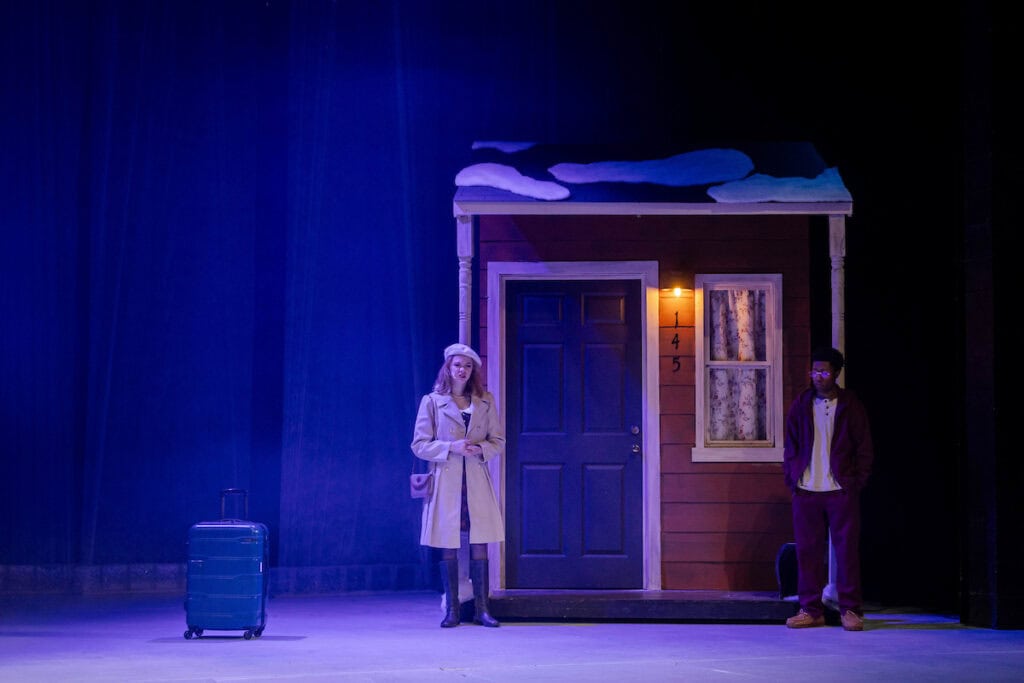 Two actors stand by a small structure onstage