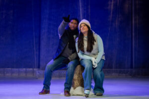 Two actors sit together onstage on a tree stump during a play rehearsal