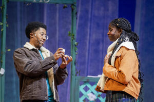 one actor holds open a ring box while another looks on, clutching her chest