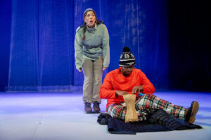 One actor stands behind another, who is sitting onstage 