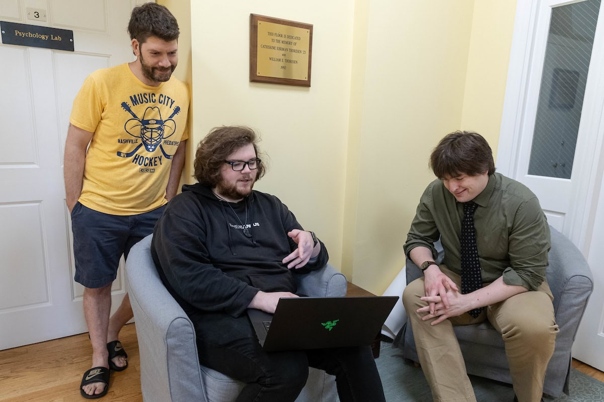 Three men--two sitting and one standing--talk, with one holding a laptop